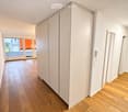 A clean, modern hallway with light wooden flooring, white walls, and a large sliding glass door leading to a balcony area with city views.