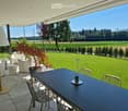 Outdoor patio with a black dining table, white chairs, white sofa, and a potted plant, overlooking a green lawn and countryside view.