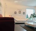 Modern living room with white walls, large windows, a white sofa, a brown leather armchair, a white coffee table, a floor lamp, and a potted plant near the window.