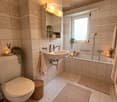 Modern bathroom with a toilet, sink, bathtub, and a large window. Features include a wicker basket, bath mats, candles, and small plants on the windowsill and shelf.