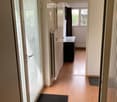 A clean, well-lit hallway with wooden flooring, white walls, and a black doormat near the entrance. A radiator is visible on the left wall, and a small black cabinet is seen at the end of the hallway.