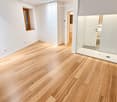 Empty room with light wooden flooring, white walls, built-in wardrobes, and a glass-enclosed shower area visible in the background.