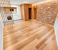 Empty room with light wooden flooring, white walls, and a brick accent wall. The room features a modern kitchen area with white cabinets and wooden accents visible in the background.
