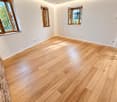 Empty room with light wooden flooring and white walls, featuring multiple windows with wooden frames and natural light streaming in.