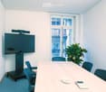 Conference room with a large screen on a rolling stand, a long wooden table surrounded by chairs, a potted plant, and a calculator on the table.