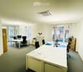 Bright office space with white desks, black chairs, computer monitors, and shelves. Natural light from large windows.