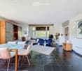 Modern living room with large windows, dark tiled floor, white sofa, blue armchairs, glass dining table, and wooden shelves.