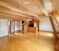 Open, spacious room with wooden flooring and exposed wooden beams on the ceiling. Features a spiral staircase leading to an upper level and a small kitchen area with cabinets and appliances.
