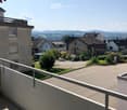 Balcony overlooking a residential area with houses, greenery, and a clear view of distant hills under a bright blue sky.