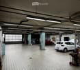 A spacious garage with tiled flooring, fluorescent lighting, and two cars, one with its hood open, along with various tools and equipment.