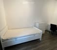 A small, neatly furnished bedroom with a single bed, a nightstand, a chair, and a desk. The room features light-colored walls and dark wooden flooring.