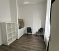 A modern, minimalist room with wooden flooring, white walls, and a small kitchen area. Two black chairs are placed near the kitchen counter, and a white shelving unit is visible on the left side.