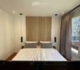 Modern bedroom with a large bed, wooden headboard, grey pillows, nightstands, and pendant lights. Large window with curtains offers a view of the city.