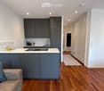 Modern kitchen with grey cabinets, white countertops, and a central island. The space features a sofa, coffee table, and a small rug in the adjacent living area. The kitchen includes a sink, faucet, and various appliances.