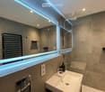 Modern bathroom with a large mirror, illuminated LED lighting, a sink with a sleek faucet, a shower area, and a radiator. The walls are tiled in a neutral tone, and a small potted plant adds a touch of greenery.