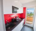 Modern kitchen with white cabinets, red accent wall, black countertop, and a view of mountains through a large window. Includes a sink, oven, microwave, toaster, and various small appliances.