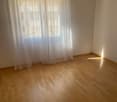 Empty room with wooden flooring and white walls, featuring a large window with sheer curtains allowing natural light to enter.