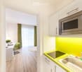 A modern, compact studio apartment with a small kitchen area featuring a microwave, sink, and faucet, and a living space with a chair, table, and a small round rug.