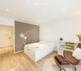 Modern bedroom with a white bed, a brown armchair, a white shelf, and a vase with flowers on the shelf.