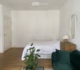 A small, modern bedroom with a bed, green sofa, nightstand with a lamp, and a decorative mirror on the wall. The room features wooden flooring and white walls.