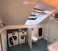 Compact bathroom with dark blue tiles, white fixtures, and a skylight window. Includes a washing machine, dishwasher, toilet, sink, and radiator.