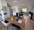 A modern, open-plan living and working space with a wooden table, laptop, chairs, kitchen area, and a comfortable seating area with a plant and screen.
