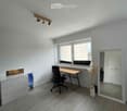 Modern, minimalist room with light grey walls, wooden floor, large window, desk, chair, mirror, and decorative vase.