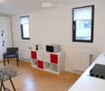 A modern, compact kitchen and living area with white walls, wooden flooring, and large windows. Features include a white cabinet with red accents, a microwave, a sink, an induction stove, a glass table, and a blue chair.