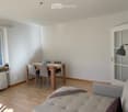 A modern, well-lit living and dining area with light wooden flooring, white walls, and large windows. Features a grey sofa, a wooden dining table with four chairs, a decorative vase, and a floor lamp near the door.