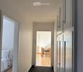 A narrow hallway with light-colored walls, carpeted floor, and white doors leading to adjacent rooms. Visible rooms include a bedroom with a white bed and a dining area with a wooden table and chairs.