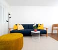 modern living room, blue sofa, yellow pillows, wooden table, potted flower, wooden floor, floor lamp, white curtains