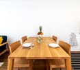 light wooden table, chairs, white walls, open to living area, bowl of fruit, glass of water, plate