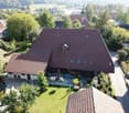 Aerial view of a large house with a brown roof, surrounded by greenery and other houses, with a driveway in front