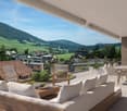 Furnished balcony with couch, chairs, table, cushions, pots, metal railing, view to village and mountains