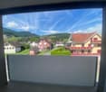 The image shows a scenic view of a small town or village nestled in a mountainous landscape. The view is framed by a large window or balcony, with a clear blue sky and fluffy white clouds overhead. The town features a mix of residential and commercial bui