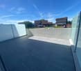 Concrete balcony with black tile flooring, overlooking a residential neighborhood with houses and a construction vehicle in the background. The sky is clear and blue with some wispy clouds.