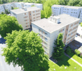 Multi-story apartment buildings with white facades, surrounded by trees and greenery, with a parking area visible in the foreground.