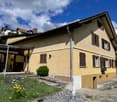 Yellow two story house with brown shutters, chimney, steps leading up to entrance, small wooden bench on porch, bicycle leaning against wall, plants, shrubs, gravel path