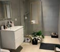 modern bathroom, grey tiles, white sink, glass enclosed shower, large mirror, potted plant, toilet, trash can