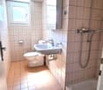 A small bathroom with tiled walls and floor, a white toilet, a white sink with a faucet, a shower head with a hose, and a shower caddy on the wall.