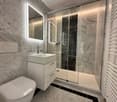 The image shows a bathroom with white porcelain tiled floors and walls, white floating vanity with sink, glass shower enclosure, white toilet bowl, and built-in illuminated mirror.