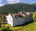 Aerial view of a modern apartment building with a brown roof, nestled in a green valley with a mountain in the background.
