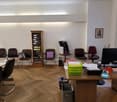 Office room with wooden floor, desks, chairs, bookshelves, computer, monitor, trash bins, picture frame, and a window.