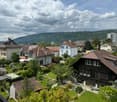 The image shows a scenic view of a residential neighborhood in a hilly area. The houses have a mix of architectural styles, including traditional Swiss-style buildings with slanted roofs. The neighborhood is surrounded by lush greenery and there are some