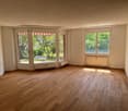 Spacious living room with large windows overlooking a garden, hardwood floors, and a bright, airy atmosphere.