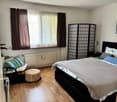 Large bedroom with wooden floor, bed, window with curtain, radiator, stool, screen, night tables, couch, desk