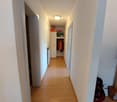 Wide hallway, white walls, wooden flooring, 4 doors, ceiling light, open door at far end, wardrobe on left, hanging clothes on right, storage box on shelf