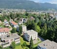 The image shows an aerial view of a town nestled in a mountainous, forested area. The town consists of a mix of residential buildings, apartment complexes, and commercial structures surrounded by lush greenery. The buildings have a variety of architectura