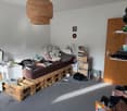 Pallet bed, wooden pallets, small table, chair, shelf, lamp, books, clutter, open door