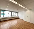 An empty room with wooden floors and white walls. Large windows with white window sill and green shutters. White built-in cabinets and a white ceiling with integrated fluorescent lighting.